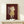 May include: Framed art print displayed on white wooden shelf featuring stylized character with red hair and yellow dress against burgundy background with window. Small wrapped gift with pine sprig sits in front of light wood frame on minimalist white wall.