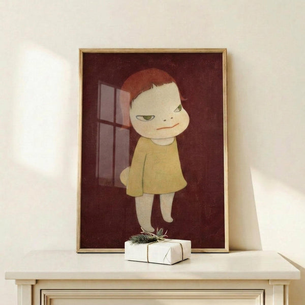 May include: Framed art print displayed on white wooden shelf featuring stylized character with red hair and yellow dress against burgundy background with window. Small wrapped gift with pine sprig sits in front of light wood frame on minimalist white wall.