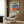 May include: Retro-style art poster in light wooden frame hanging on dark wood-paneled wall above beige upholstered armchair with geometric patterned pillow. Artwork shows collage-style composition with blue swimming pool water, orange color block, woman wearing headphones, and rocky beach elements. Modern brass floor lamp and dark round side table with open book complete the contemporary interior setting.