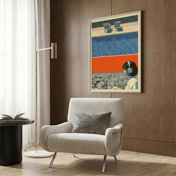 May include: Retro-style art poster in light wooden frame hanging on dark wood-paneled wall above beige upholstered armchair with geometric patterned pillow. Artwork shows collage-style composition with blue swimming pool water, orange color block, woman wearing headphones, and rocky beach elements. Modern brass floor lamp and dark round side table with open book complete the contemporary interior setting.