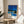May include: Framed art print displayed on wooden side table next to beige upholstered chair. The poster shows a modernist poolside scene with bright blue swimming pool, white mid-century modern house, palm trees against deep blue sky, and colorful dot pattern at bottom. Natural wood frame complements the minimalist room setting with white walls and soft natural lighting.