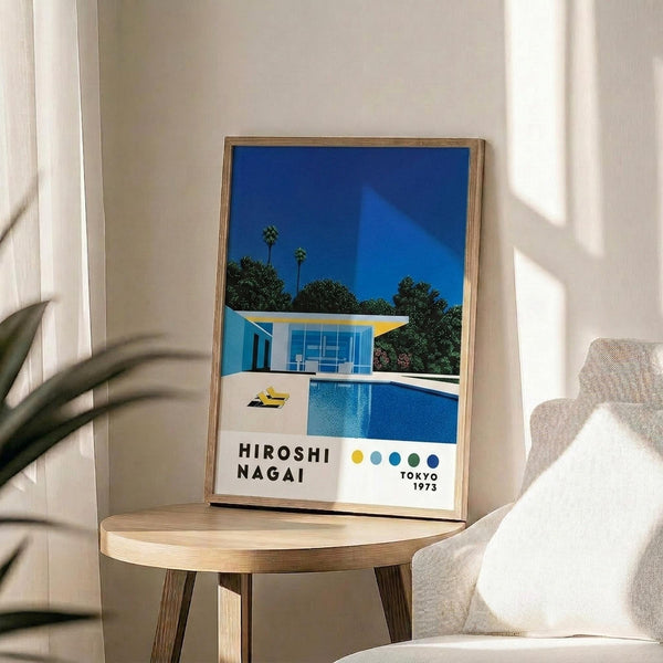 May include: Framed art print displayed on wooden side table next to beige upholstered chair. The poster shows a modernist poolside scene with bright blue swimming pool, white mid-century modern house, palm trees against deep blue sky, and colorful dot pattern at bottom. Natural wood frame complements the minimalist room setting with white walls and soft natural lighting.