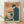 May include: Framed vintage-style poster featuring a person in dark clothing looking at a retro beige computer with CRT monitor and keyboard. The artwork uses orange, teal, and cream colors in a nostalgic 1980s aesthetic with Japanese text at the top. The wooden frame sits on light wood flooring against a white wall with dramatic shadows from window blinds creating striped patterns across the scene.