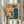 May include: Person in cream sweater holding framed vintage-style poster featuring man at retro computer, orange and teal color scheme with Japanese text, alongside dried pampas grass stems against soft natural lighting and neutral curtained background