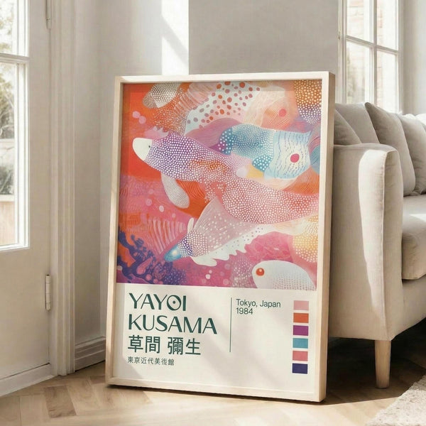 May include: Framed art print leaning against white wall next to beige sofa in bright living room. Poster features abstract colorful design with flowing organic shapes in coral, pink, blue and purple tones with dotted patterns. White frame with exhibition-style text below artwork showing "YAYOI KUSAMA" and "Tokyo, Japan 1984" with Japanese characters and color swatches. Natural sunlight streams through tall windows.