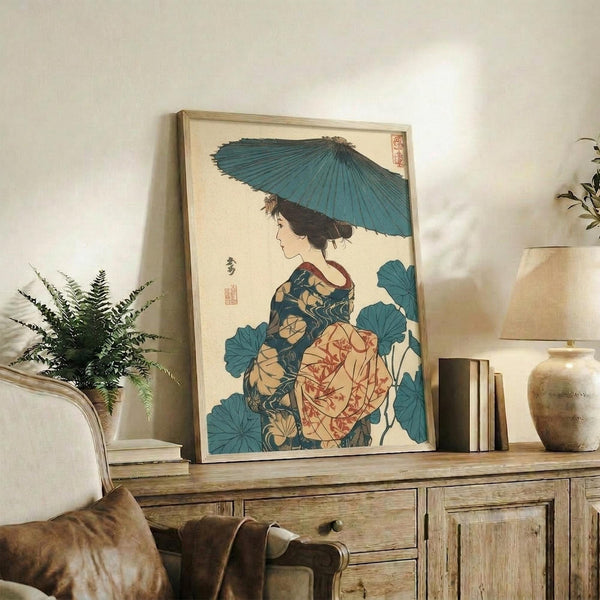 May include: Framed Japanese woodblock print artwork featuring a woman in colorful kimono holding a blue parasol, surrounded by large teal lotus leaves. The vintage-style print is displayed on a rustic wooden sideboard next to a fern plant, ceramic vase, and books in a neutral-toned living room setting with beige chair visible.