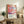 May include: Colorful abstract poster with flowing organic shapes in coral, pink, and blue tones featuring dotted patterns, displayed in light wood frame on rustic wooden sideboard alongside fern plant, books, ceramic vase, and beige upholstered chair in bright modern living room