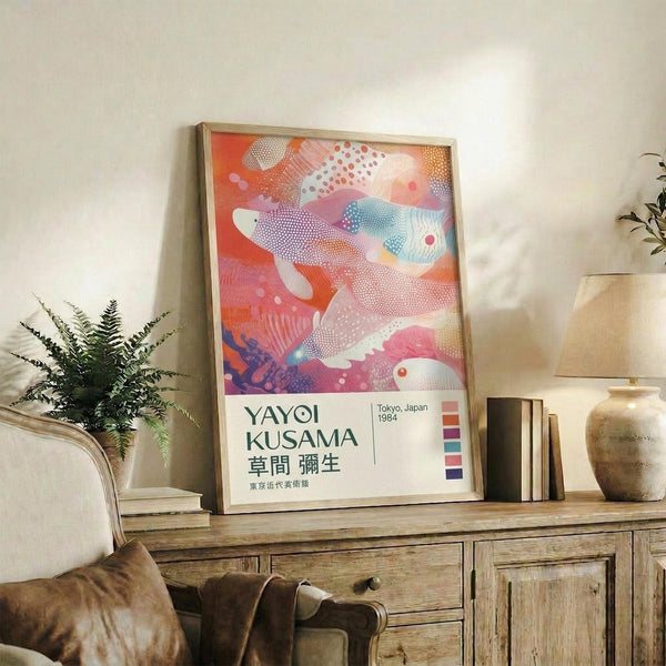 May include: Colorful abstract poster with flowing organic shapes in coral, pink, and blue tones featuring dotted patterns, displayed in light wood frame on rustic wooden sideboard alongside fern plant, books, ceramic vase, and beige upholstered chair in bright modern living room
