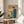May include: Framed vintage-style poster with Japanese text and retro computer illustration in teal and orange colors, displayed on round wooden side table next to beige upholstered chair. Sunlight streams through white curtains creating soft shadows, with palm plant leaves visible in corner of bright, minimalist room setting.