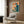 May include: Framed retro-style poster with orange and teal colors showing figure at vintage computer, displayed on wood paneled wall above beige upholstered armchair with geometric patterned pillow, brass floor lamp with white marble base, and dark round side table with open book in modern living room setting