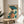 May include: Framed Japanese woodblock print artwork displayed on wooden round side table next to beige upholstered chair. Print shows woman in traditional kimono with floral patterns holding blue-green parasol, surrounded by large teal leaves. Warm natural lighting creates soft shadows on cream walls. Plant leaves visible in corner of minimalist room setting.