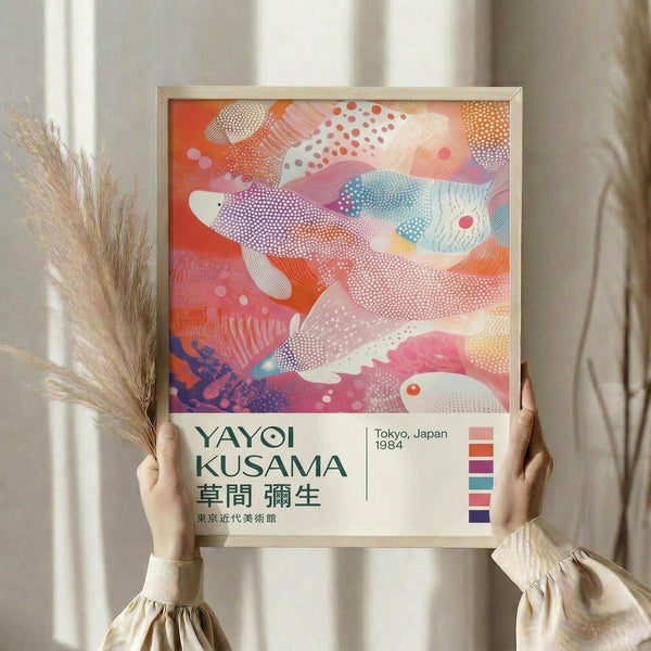 May include: Person holding framed art print featuring abstract fish shapes in vibrant coral, pink, blue and orange colors with dotted patterns. Poster displays "YAYOI KUSAMA" text and "Tokyo, Japan 1984" with Japanese characters below. Natural wood frame against neutral curtained background with dried wheat stems visible on left side.