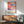 May include: Framed colorful abstract art poster featuring flowing organic shapes in pink, orange, blue and purple tones with dotted patterns, displayed above a mid-century modern wooden credenza with white ceramic lamp, next to sage green upholstered armchair in bright sunlit room with white walls.