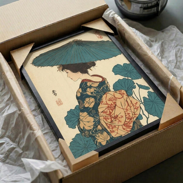 Framed Japanese woodblock print featuring a woman in traditional kimono holding a teal umbrella, surrounded by large green lotus leaves. The artwork shows muted earth tones with coral and teal accents on aged paper. The black frame sits in an open cardboard shipping box with protective wrapping materials and rolled tubes visible around it.