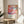 May include: Abstract colorful poster featuring flowing organic shapes in vibrant pink, orange, blue and purple with dotted patterns, displayed in thin gold frame on round wooden side table next to beige upholstered chair. Natural sunlight streams across neutral wall, with green plant leaves visible in corner creating soft shadows.