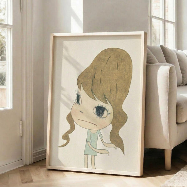 May include: Framed art print featuring a cartoon-style illustration of a young girl with wavy golden-brown hair and large blue eyes, wearing a light green dress. The artwork has a soft, hand-drawn aesthetic on cream-colored paper within a light wood frame. The print is displayed leaning against a beige upholstered sofa in a bright, airy living room with white walls and large windows casting natural light across polished floors.