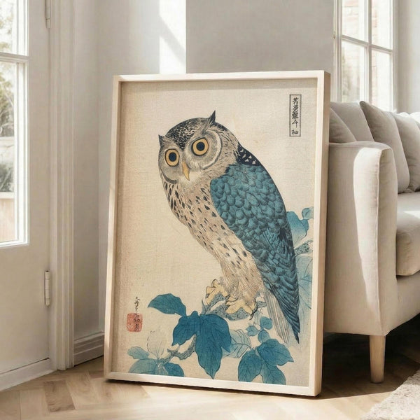 May include: Japanese woodblock print style artwork of an owl with large golden eyes perched on a branch with blue-green foliage, displayed in a light wooden frame leaning against a white wall beside a beige upholstered chair in a bright room with large windows