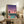 May include: Framed poster showing a retro-style illustration of a rooftop swimming pool overlooking a city skyline at sunset, with palm trees silhouetted against a purple-pink gradient sky and an airplane in the distance. The artwork sits on a rustic wooden console table alongside books, a ceramic vase, and a potted fern, with a cream upholstered chair visible in the foreground.
