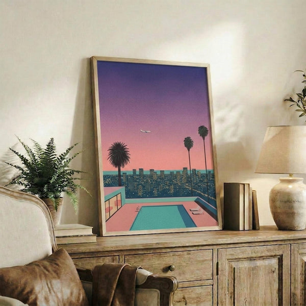 May include: Framed poster showing a retro-style illustration of a rooftop swimming pool overlooking a city skyline at sunset, with palm trees silhouetted against a purple-pink gradient sky and an airplane in the distance. The artwork sits on a rustic wooden console table alongside books, a ceramic vase, and a potted fern, with a cream upholstered chair visible in the foreground.
