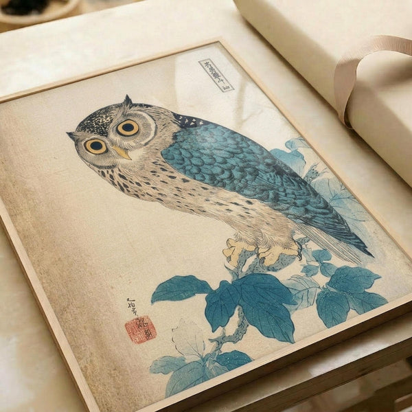 Japanese woodblock print featuring an owl perched on a flowering branch with blue-green leaves. The owl has detailed brown and cream feathering with distinctive large yellow eyes and intricate line work typical of traditional ukiyo-e art. Red artist seal visible in lower left corner. Print is matted in cream-colored mat and displayed on white surface with rolled papers nearby, showing vintage paper texture and authentic aged appearance.