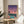May include: Framed poster displaying a retro sunset cityscape with pink and purple sky, palm trees, turquoise swimming pool, and city lights below. Artwork sits on round wooden side table next to beige upholstered chair with white pillow. Soft natural lighting filters through window creating shadows on cream wall. Green plant leaves visible in corner.