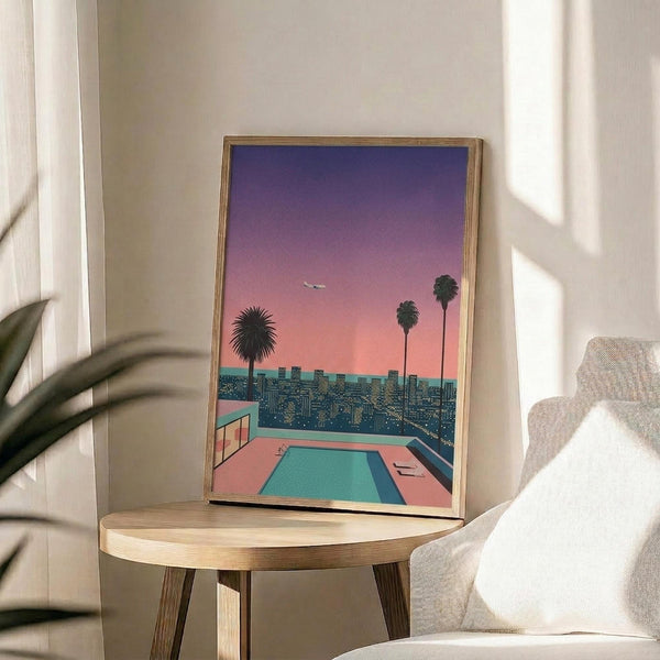 May include: Framed poster displaying a retro sunset cityscape with pink and purple sky, palm trees, turquoise swimming pool, and city lights below. Artwork sits on round wooden side table next to beige upholstered chair with white pillow. Soft natural lighting filters through window creating shadows on cream wall. Green plant leaves visible in corner.