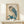 May include: Framed Japanese woodblock print of an owl with large yellow eyes perched on blue-green foliage, displayed on light wood furniture against white wall. Print shows detailed feather textures in brown, gray and teal colors with traditional Asian calligraphy. Small white wrapped gift sits beside the wooden frame on the shelf surface.