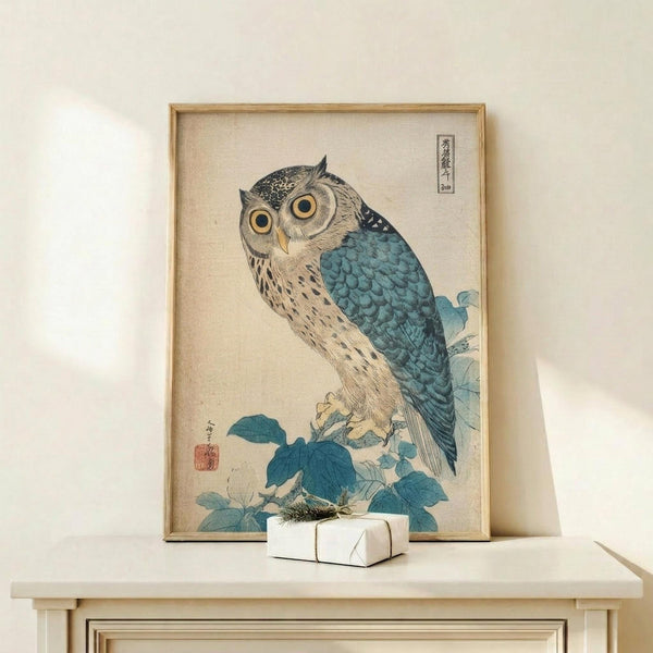 May include: Framed Japanese woodblock print of an owl with large yellow eyes perched on blue-green foliage, displayed on light wood furniture against white wall. Print shows detailed feather textures in brown, gray and teal colors with traditional Asian calligraphy. Small white wrapped gift sits beside the wooden frame on the shelf surface.