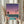 May include: Person in cream ruffled sleeves holding framed poster print in light wooden frame. Artwork shows retro-style illustration with gradient purple-to-pink sunset sky, turquoise swimming pool in foreground, city skyline with lit buildings, three palm tree silhouettes, and small airplane. Dried pampas grass visible on left side against soft beige curtains and white window frame.