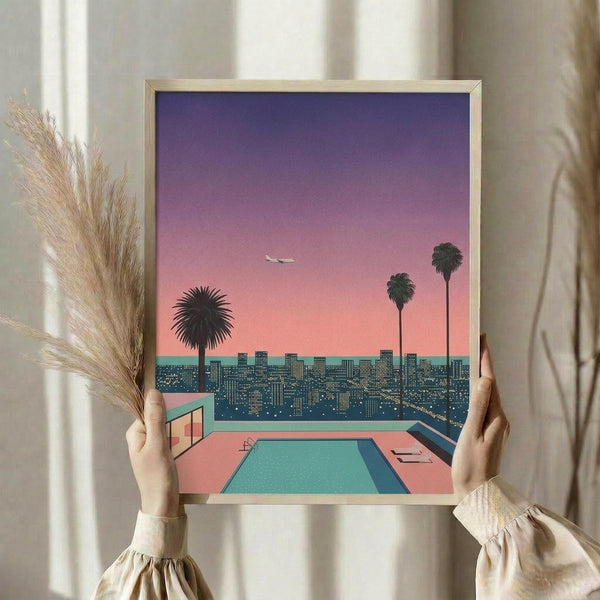 May include: Person in cream ruffled sleeves holding framed poster print in light wooden frame. Artwork shows retro-style illustration with gradient purple-to-pink sunset sky, turquoise swimming pool in foreground, city skyline with lit buildings, three palm tree silhouettes, and small airplane. Dried pampas grass visible on left side against soft beige curtains and white window frame.