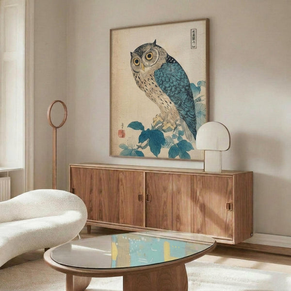 May include: Japanese woodblock print of an owl with blue wings perched on flowering branch, displayed in light wood frame above mid-century walnut credenza. Modern living room setting with cream curved chair, oval glass coffee table, and brass floor lamp. Neutral walls with natural lighting creating shadows.