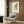 May include: Framed art print featuring a cartoon-style girl with voluminous blonde beehive hair and large eyes, wearing a light green dress, displayed on wood-paneled wall above a cream upholstered armchair with geometric patterned pillow, beside a brass floor lamp and dark round side table with open book