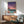 May include: Retro-style artwork in wooden frame showing sunset cityscape with swimming pool, palm trees, and purple-pink sky, displayed above mid-century modern wooden credenza with white ceramic lamp, next to sage green upholstered armchair with wooden arms in minimalist room with white walls and natural lighting.