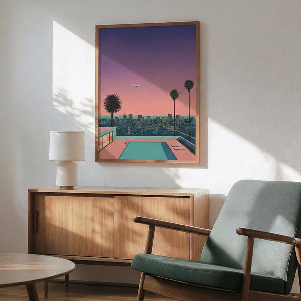 May include: Retro-style artwork in wooden frame showing sunset cityscape with swimming pool, palm trees, and purple-pink sky, displayed above mid-century modern wooden credenza with white ceramic lamp, next to sage green upholstered armchair with wooden arms in minimalist room with white walls and natural lighting.