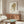 May include: Modern minimalist living room featuring framed wall art of a cartoon-style girl with blonde beehive hair and large eyes wearing a mint green dress. The artwork hangs above a mid-century walnut credenza with brass handles, flanked by a copper floor lamp and white dome table lamp. A white curved lounge chair and oval glass coffee table complete the neutral-toned space with natural lighting.