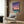 May include: Framed poster displaying a retro-style cityscape with swimming pool in foreground, palm trees, and gradient sunset sky in purple and pink tones. Shown mounted on wood-paneled wall above beige upholstered armchair with geometric patterned pillow. Modern floor lamp with brass finish and round side table with open book visible in styled room setting with white curtains.