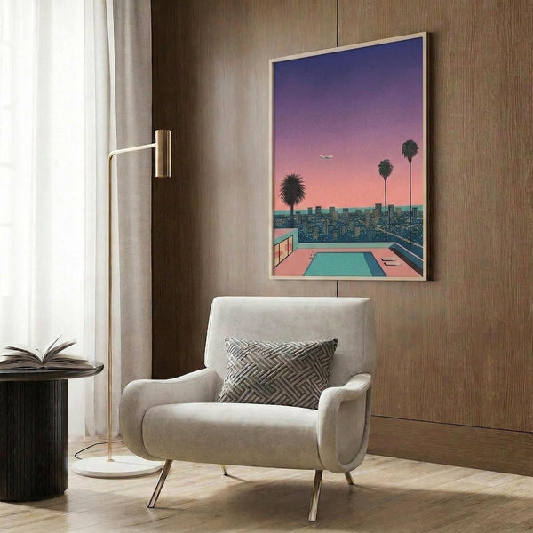 May include: Framed poster displaying a retro-style cityscape with swimming pool in foreground, palm trees, and gradient sunset sky in purple and pink tones. Shown mounted on wood-paneled wall above beige upholstered armchair with geometric patterned pillow. Modern floor lamp with brass finish and round side table with open book visible in styled room setting with white curtains.