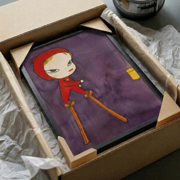 May include: Framed artwork featuring a cartoon-style child character with blonde hair wearing a red hooded coat, skiing on orange skis against a purple background with a small yellow window. The black-framed print sits in an open cardboard shipping box surrounded by white tissue paper, with wooden corner protectors visible. A glass bowl appears in the background on a white surface.