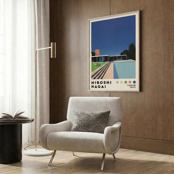 May include: Framed poster featuring modernist pool scene with turquoise water, wooden deck, glass house, and deep blue sky, displayed on wood-paneled wall above beige upholstered armchair with geometric patterned pillow. Gold floor lamp and dark round side table with open book visible in contemporary interior setting with warm wood tones and natural lighting from window.