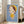 May include: Framed poster print leaning against beige armchair in bright living room with white walls and windows. Artwork shows stylized child character with bob haircut wearing yellow dress and headband on blue background. Light wood frame, approximately 2-3 feet tall, displayed on hardwood floors near gray upholstered furniture.