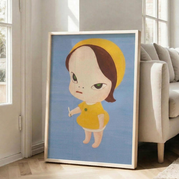 May include: Framed poster print leaning against beige armchair in bright living room with white walls and windows. Artwork shows stylized child character with bob haircut wearing yellow dress and headband on blue background. Light wood frame, approximately 2-3 feet tall, displayed on hardwood floors near gray upholstered furniture.