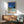 May include: Framed poster artwork displayed above wooden credenza in modern living room with mint green armchair. Poster shows modernist pool scene with sleek house, swimming pool, and wooden deck under deep blue sky. Features bold geometric shapes in blues, greens, and warm tones with "HIROSHI NAGAI" and "TOKYO 1973" text. Natural wood frame complements mid-century furniture styling. Sunlight casts diagonal shadows across white wall.