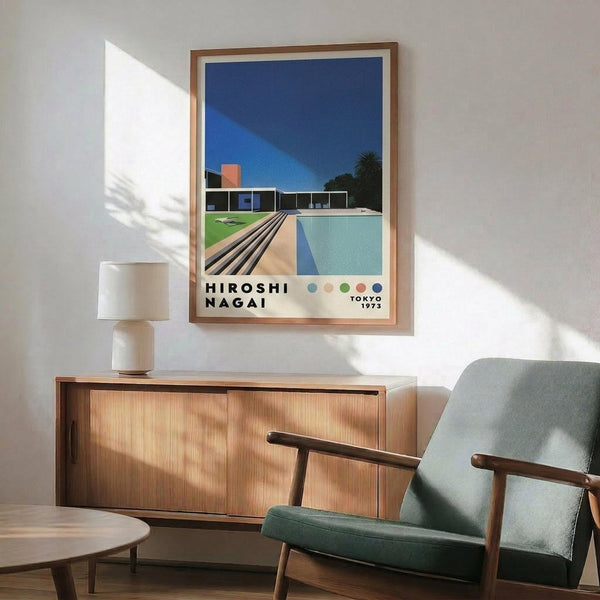 May include: Framed poster artwork displayed above wooden credenza in modern living room with mint green armchair. Poster shows modernist pool scene with sleek house, swimming pool, and wooden deck under deep blue sky. Features bold geometric shapes in blues, greens, and warm tones with "HIROSHI NAGAI" and "TOKYO 1973" text. Natural wood frame complements mid-century furniture styling. Sunlight casts diagonal shadows across white wall.