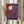 May include: Person in beige clothing holding a framed art print featuring a stylized child character in red hooded outfit and wooden stilts against purple background, with dried pampas grass visible to the left, neutral home interior setting