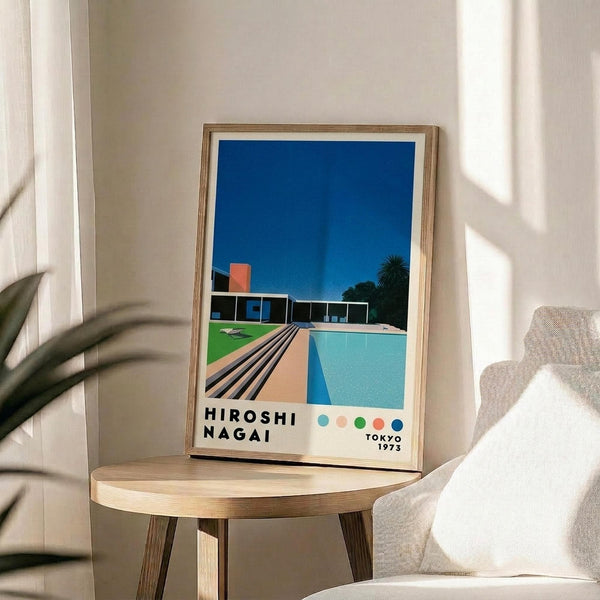 May include: A framed art print displaying a modernist poolside scene with geometric architecture, bright blue sky, turquoise swimming pool, and orange accent building. The poster features "HIROSHI NAGAI" and "TOKYO 1973" text with colorful dots below. The wooden-framed print sits on a round natural wood side table next to a beige upholstered chair, with plant shadows casting across white walls in soft natural lighting.