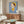 May include: Modern living room interior featuring a framed poster of a stylized cartoon child character with brown bob haircut, green eyes, wearing yellow dress and headband against blue background. The artwork hangs above a mid-century walnut credenza with sliding doors. Room includes white curved lounge chair, brass floor lamp, white table lamp, oval glass coffee table, and cream textured area rug on light hardwood floors.