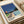 May include: Framed poster print displayed on white coffee table next to beige upholstered chair. Artwork shows stylized mid-century modern pool scene with geometric swimming pool, modernist house, palm tree, and deep blue sky. Features vintage-inspired illustration style with bold colors including turquoise water, coral and green accents. Text reads "Hiroshi Nagai" and "Tokyo 1973" with colorful dots. Natural wood frame complements minimalist home decor setting.