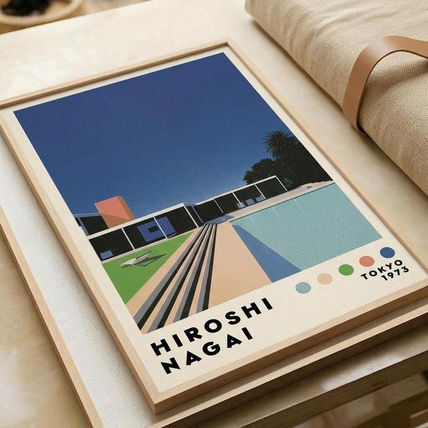 May include: Framed poster print displayed on white coffee table next to beige upholstered chair. Artwork shows stylized mid-century modern pool scene with geometric swimming pool, modernist house, palm tree, and deep blue sky. Features vintage-inspired illustration style with bold colors including turquoise water, coral and green accents. Text reads "Hiroshi Nagai" and "Tokyo 1973" with colorful dots. Natural wood frame complements minimalist home decor setting.