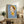 May include: Framed art print featuring a stylized illustration of a young child with brown and yellow hair wearing a bright yellow outfit, holding a small white object, set against a soft blue background. The wooden-framed artwork leans against white wall on rustic wooden sideboard, surrounded by potted plants, stacked books, ceramic vase, and brown leather furniture in warm, naturally-lit living room setting.