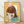 May include: Framed artwork featuring a cartoon-style illustration of a pale child with brown bowl-cut hair and large yellow-green eyes on yellow background. Mixed media piece incorporates vintage documents and forms as collage elements. Natural wood frame sits on light wooden floor against white wall with soft shadows.